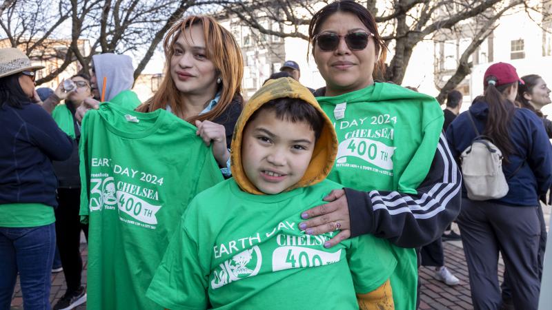 A family of three volunteers wearing green "earth day 2024" t-shirts smiling during a community event on a sunny street.