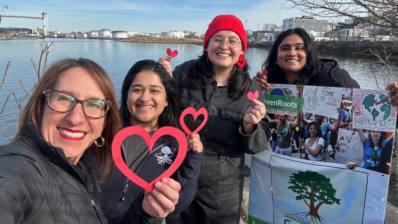 Four smiling individuals holding heart shapes and a GreenRoots sign by a riverside.