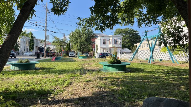 Park located at 212 Congress Ave, in Chelsea, MA. There is a swing and planters as well as shade from trees. The sky is bright blue.