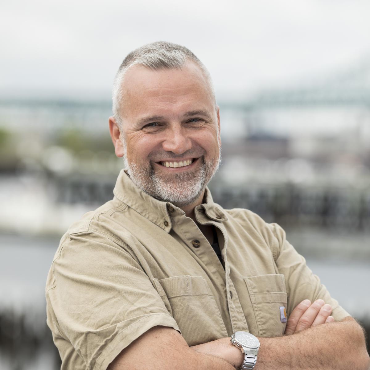 A man with salt and pepper hair with a tan shirt standing in front of the Chelsea Creek.