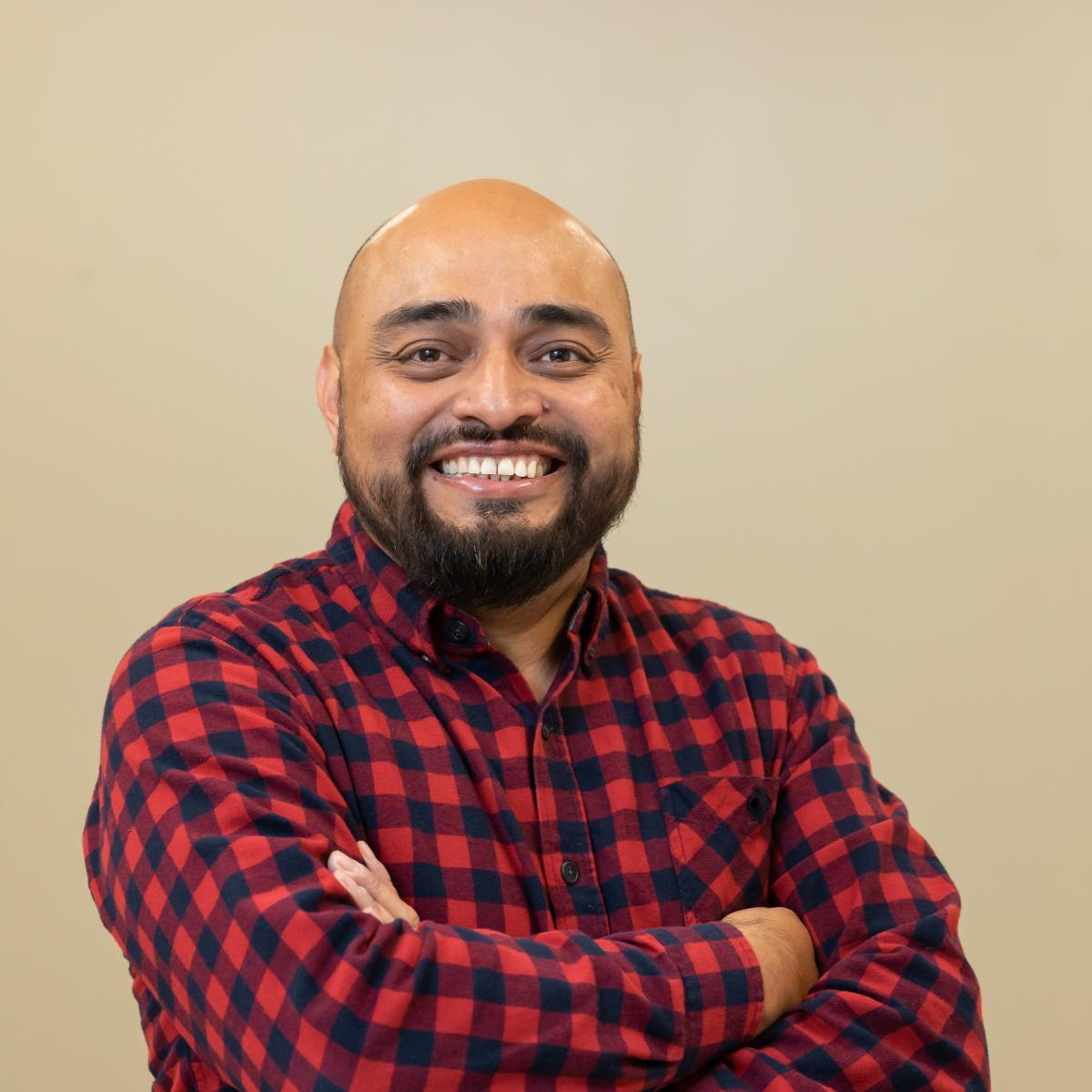 A photo of a man with a red and black plaid shirt and arms crossed standing in front of a tan wall with a dark beard.