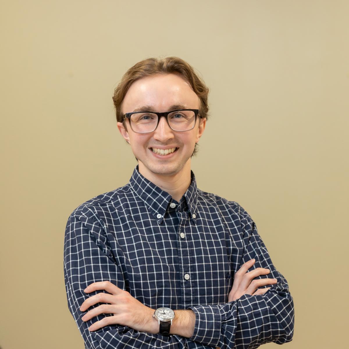 A photo of a young man with with blonde hair and wearing glasses with black frames standing in front of a tan wall with his arms crossed.