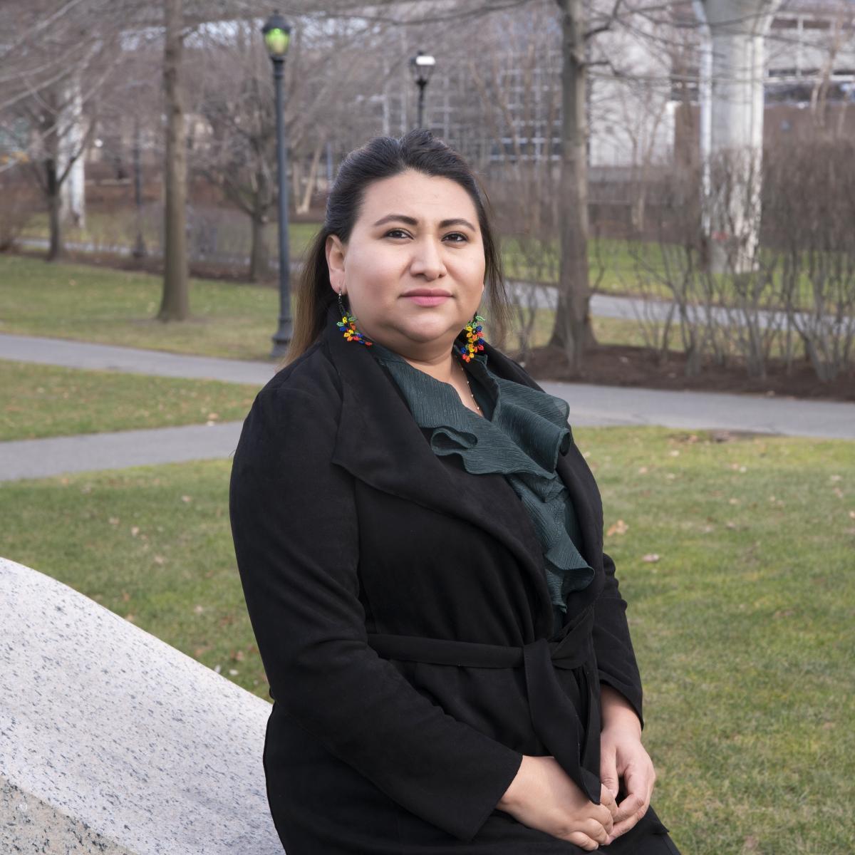 A woman with a black blazer and long earrings standing in a green area.