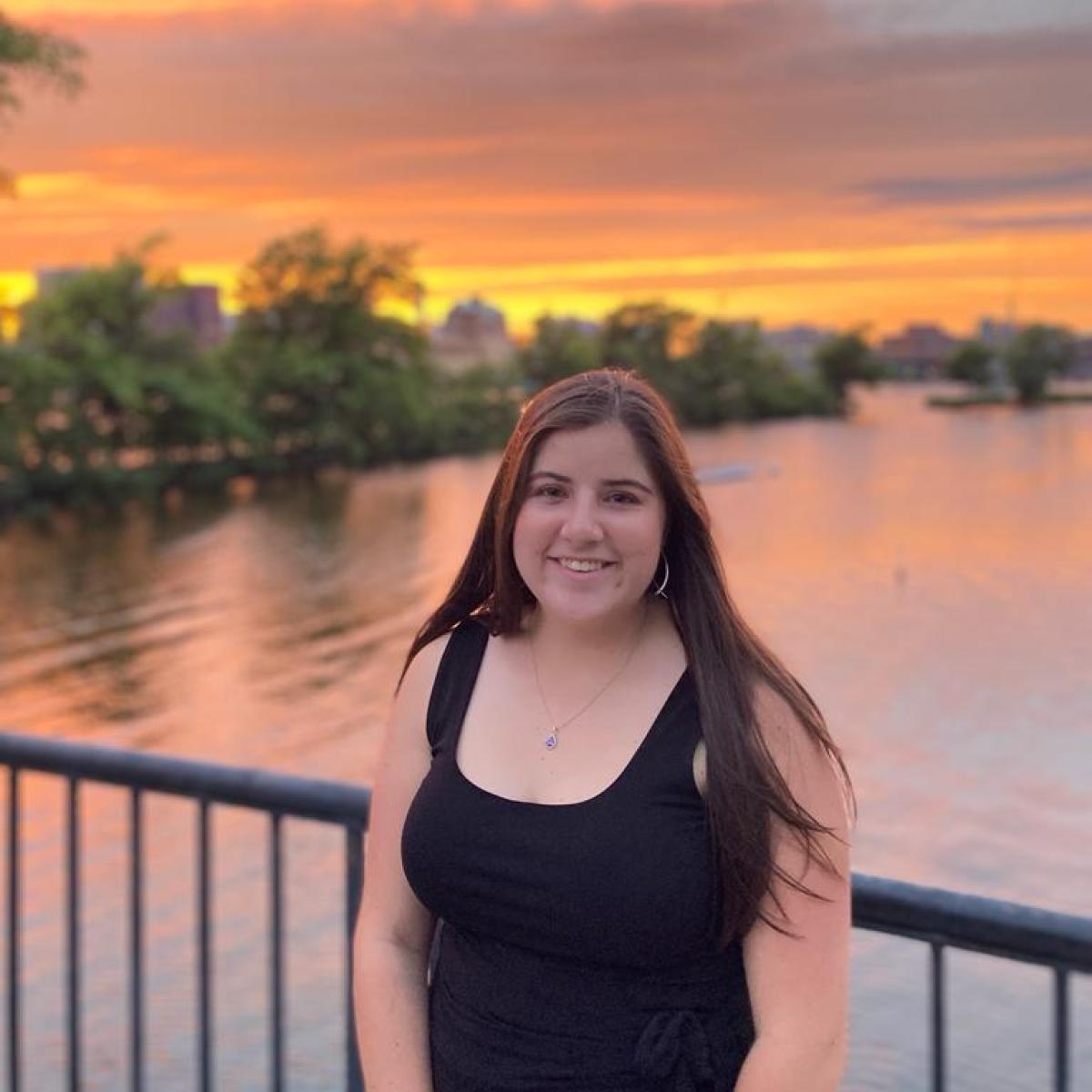 A woman with a black dress and long hair standing in front of the sunset by the water.