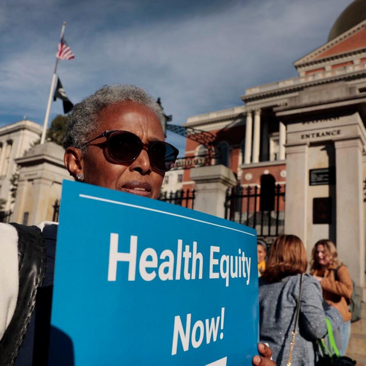 Black woman wearing sunglasses holding a sign that reads "Health Equity Now!" in front of the Massachusetts State House.