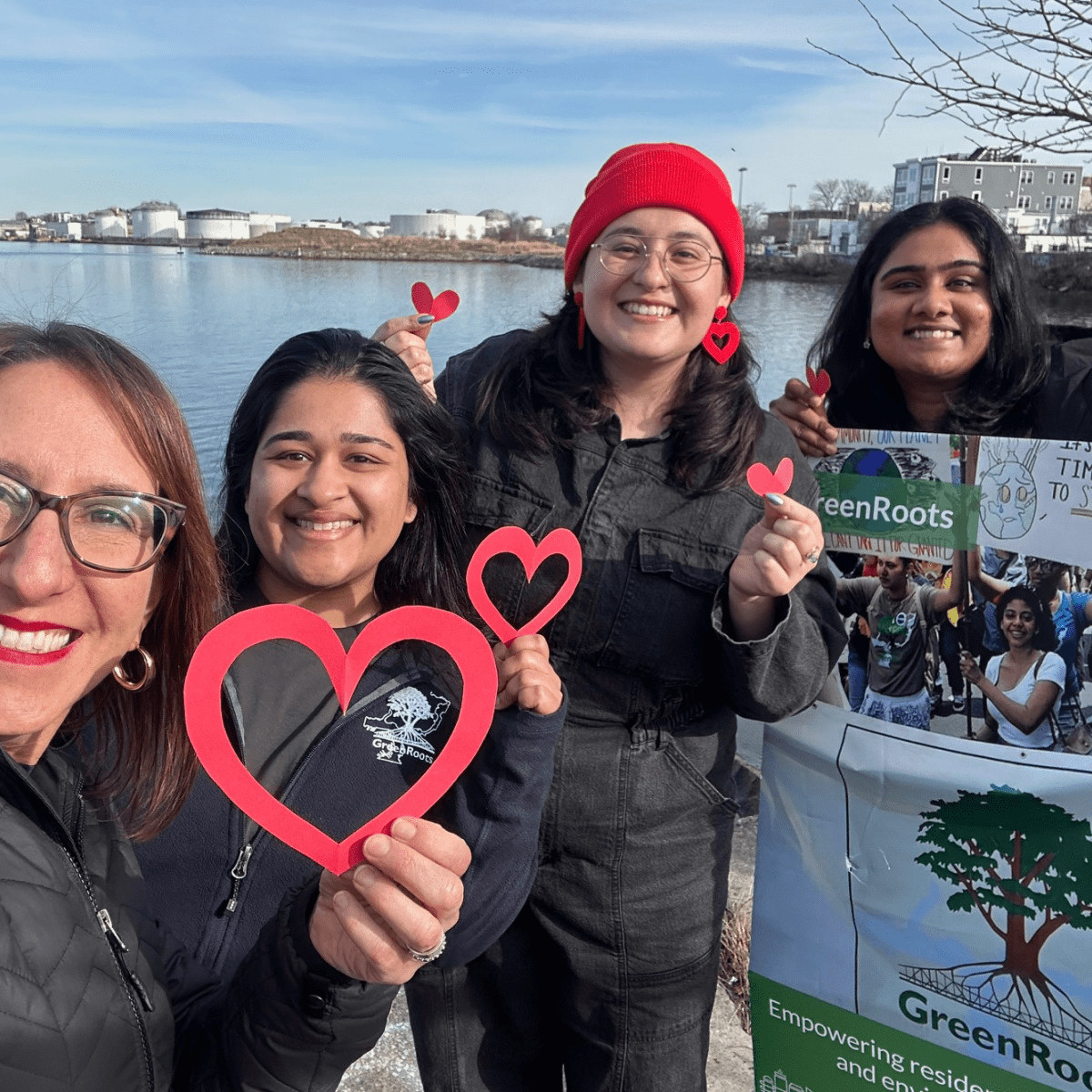 Four smiling individuals holding heart shapes and a GreenRoots sign by a riverside.