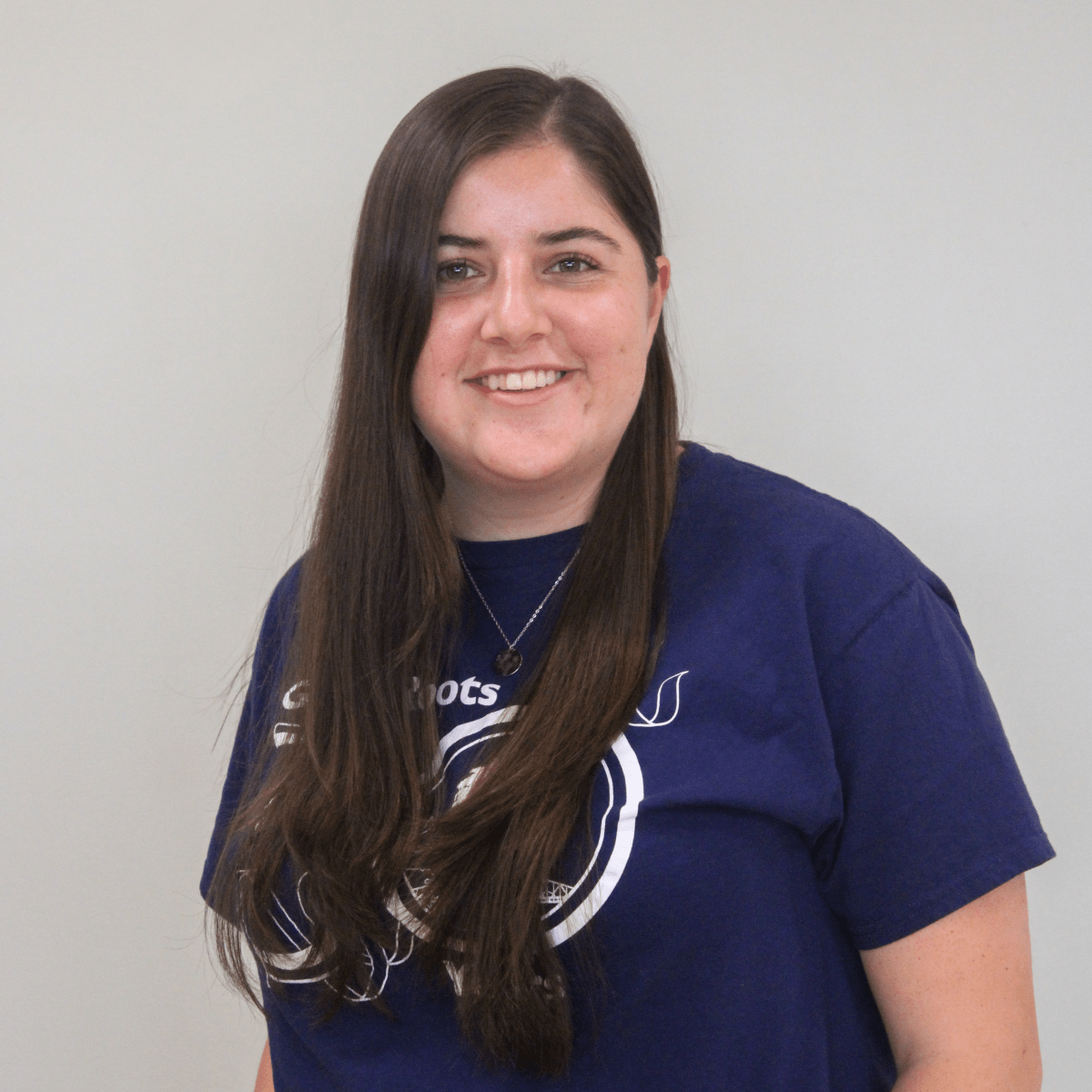 A woman in a blue shirt and necklace standing against a light wall.