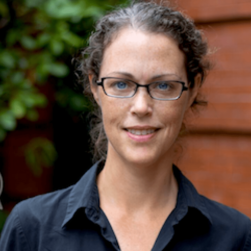 A middle-aged woman with her hair parted in the middle and a black top and blazer against a tree a wooden wall.