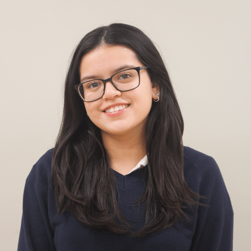 Smiling teenage girl wearing glasses and a navy blue sweater against a white wall. 