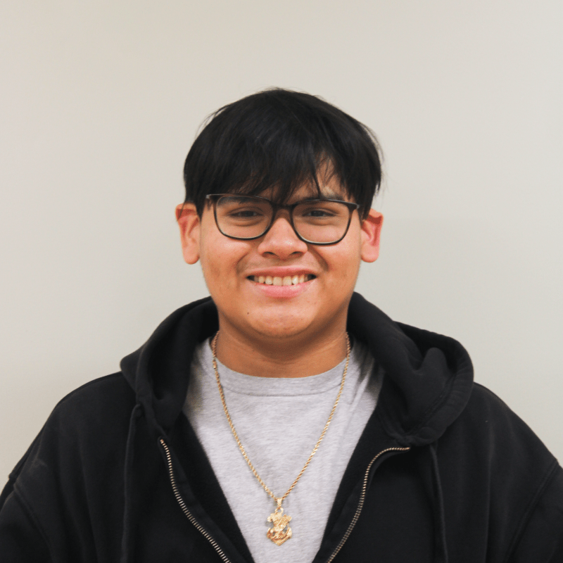 Smiling teenage boy wearing glasses, a gray shirt and black hoodie against a white wall.