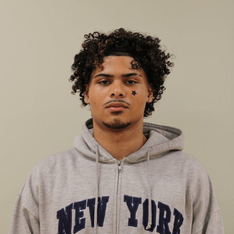 A male teenager with curly hair and a star sticker on their face is wearing a gray "New York" hoodie, standing against a plain beige background.
